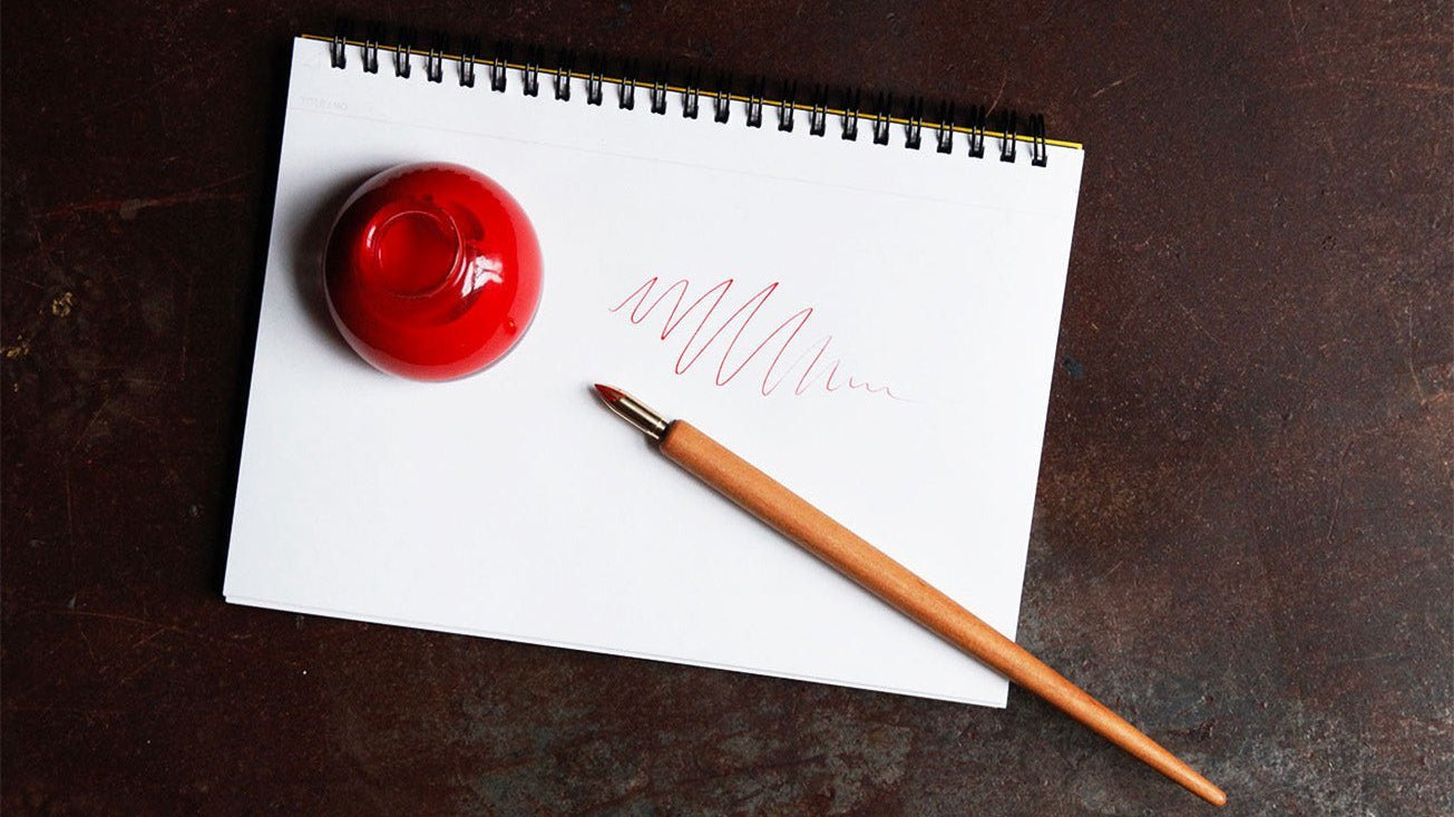 Kakimori wooden nib holder next to bottle of red ink on pad of paper on dark background