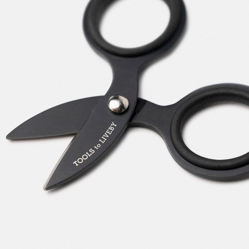 Close up of small black steel scissors with 'Tools to Liveby' branding on blade on white surface