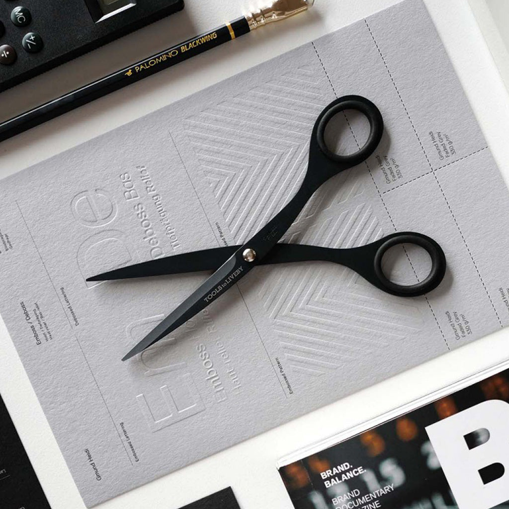 Black stainless steel scissors with 'TOOLS to LIVEBY' branding on blades on a gray sheet of paper.