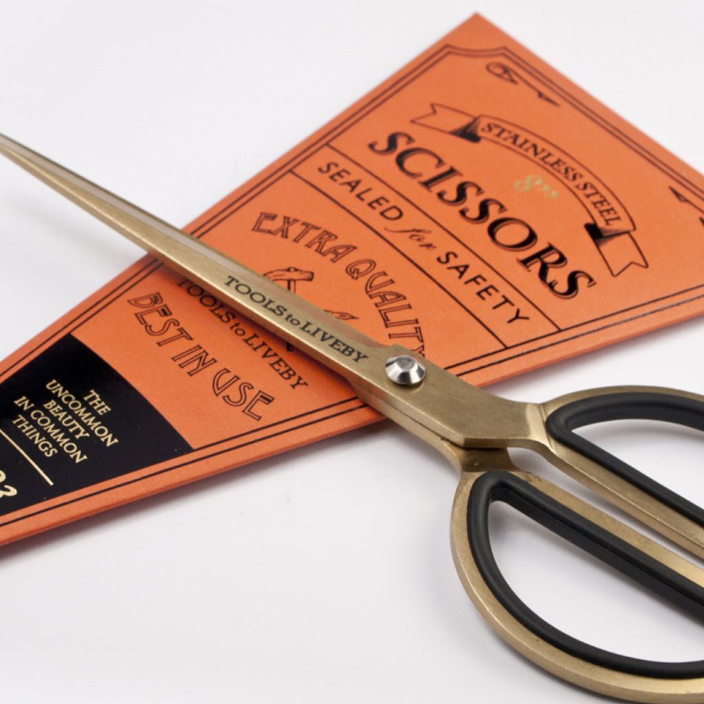 Gold-toned scissors with black rubberized finger loops on orange packaging