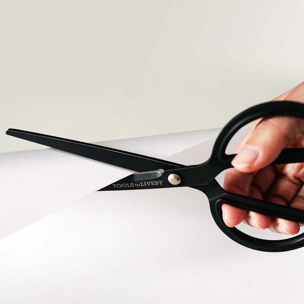 A close-up of a hand holding a pair of black scissors with the brand name 'TOOLS to LIVEBY' printed on the blade, cutting a sheet of white paper against a light neutral background.