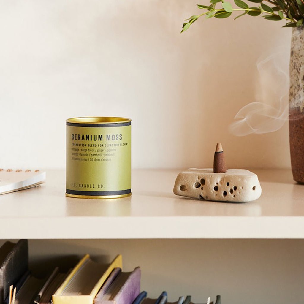 Canister of incense cones labeled 'Geranium Moss' next to a ceramic incense burner with smoke on a shelf.