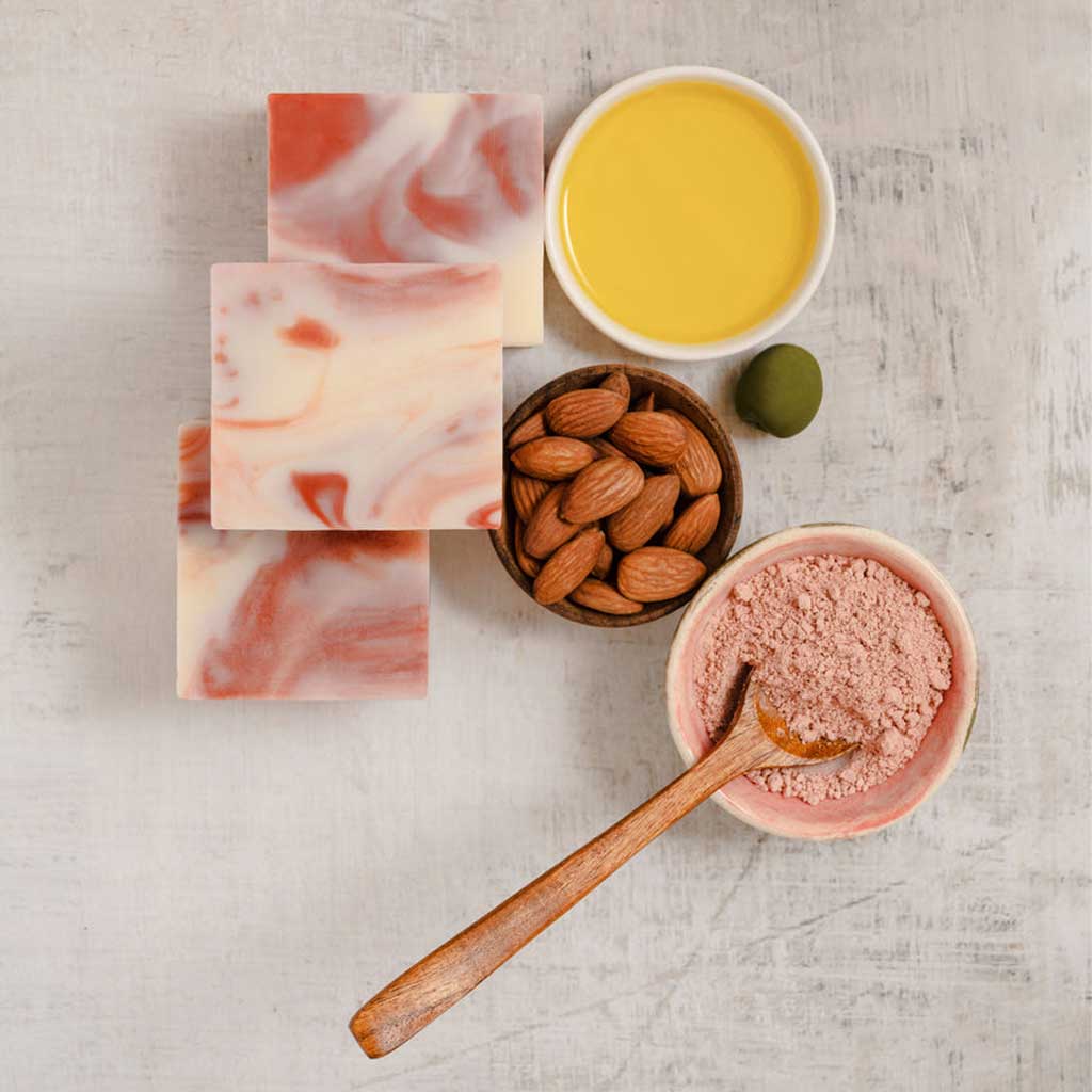 Marble-patterned SallyeAnder almond goat milk soap bars, a bowl of olive oil, a bowl of pink french clay, and a bowl of almonds on a light gray surface.