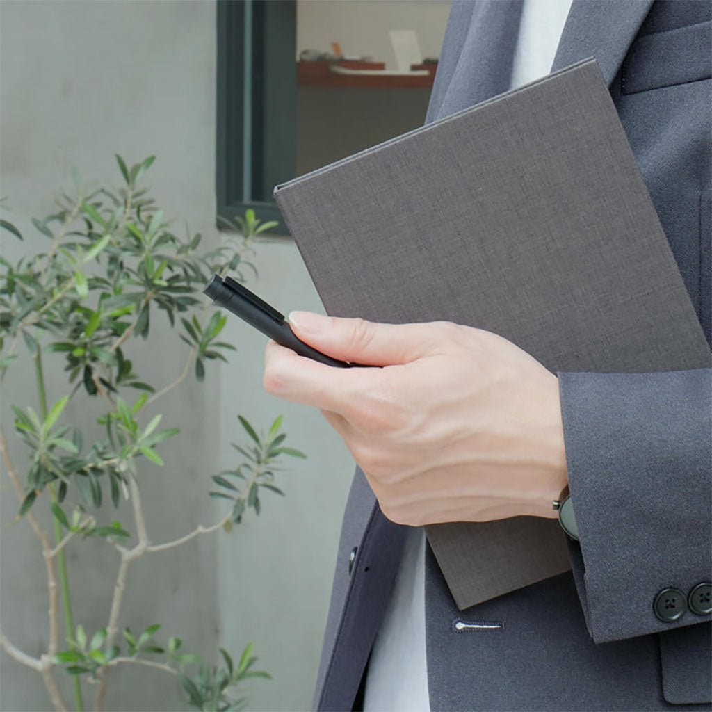Person holding a gray notebook and pen outdoors with a plant in the background
