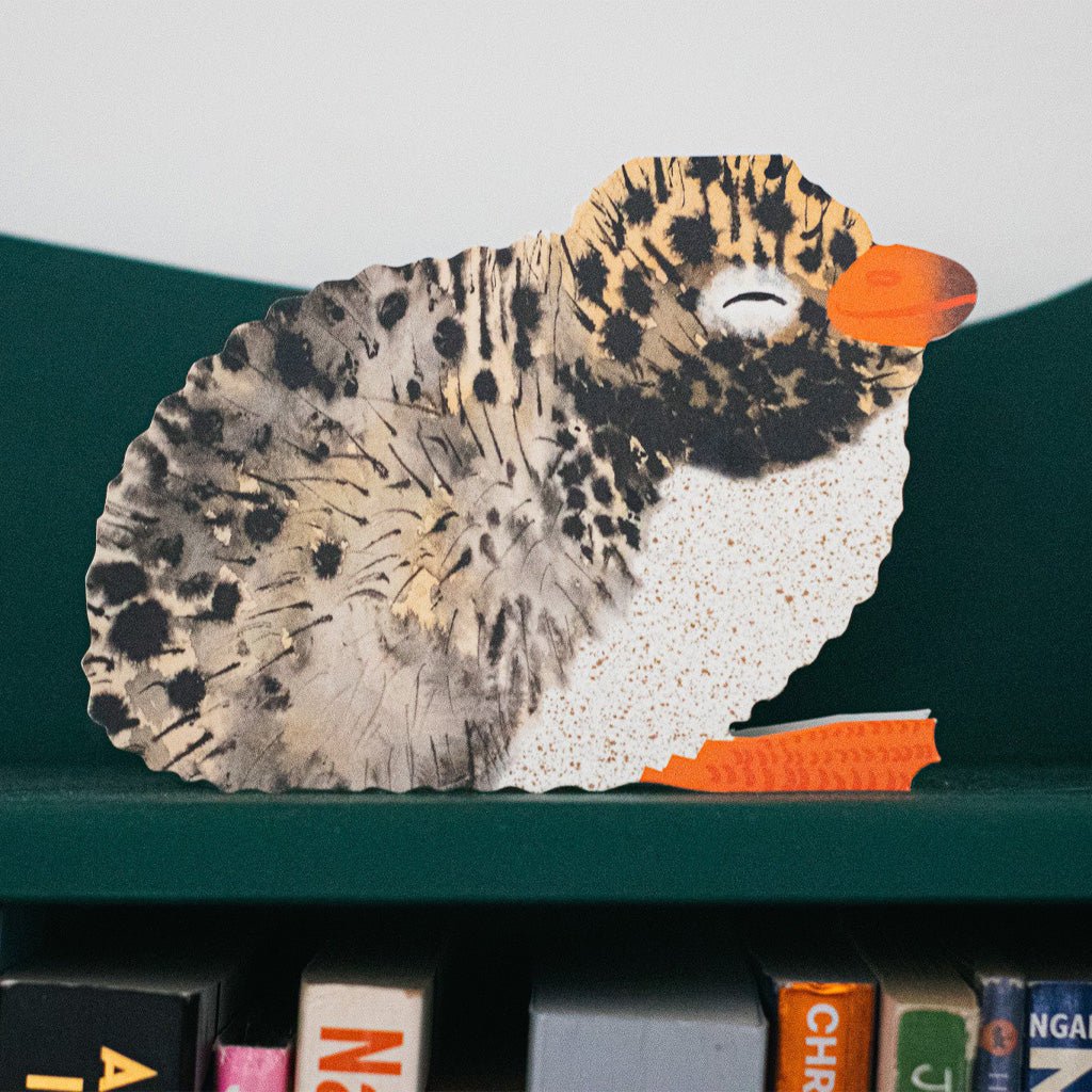 A decorative cutout of a arctic tern with a spotted black-and-white body, orange beak, and orange feet, displayed on a dark green shelf above books.