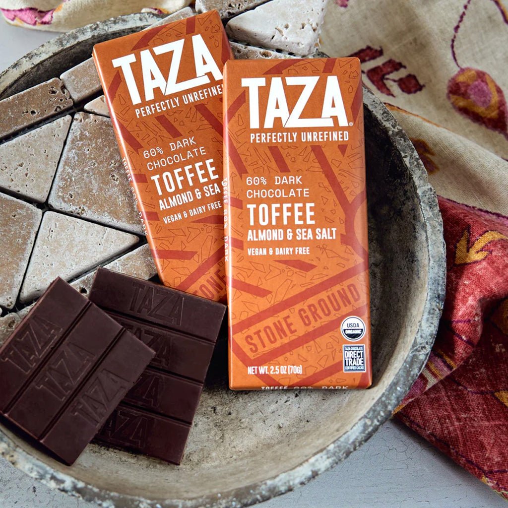Two TAZA chocolate bars with toffee flavor next to packaging on a stone surface.