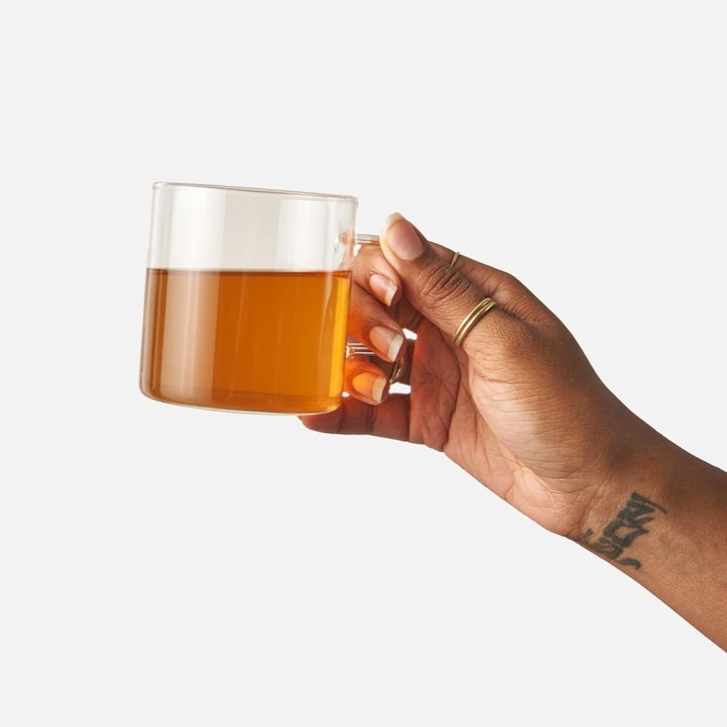 A hand holding a clear glass mug filled with amber tea against a plain light gray background.