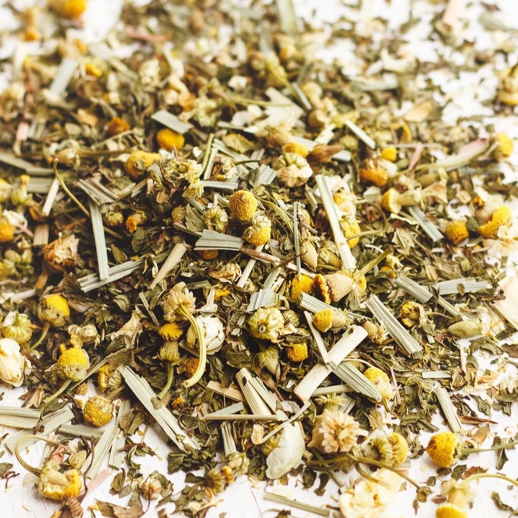 A close-up of loose-leaf chamomile tea blend, featuring dried green leaves, pale stems, and small yellow chamomile flowers scattered on a light background.