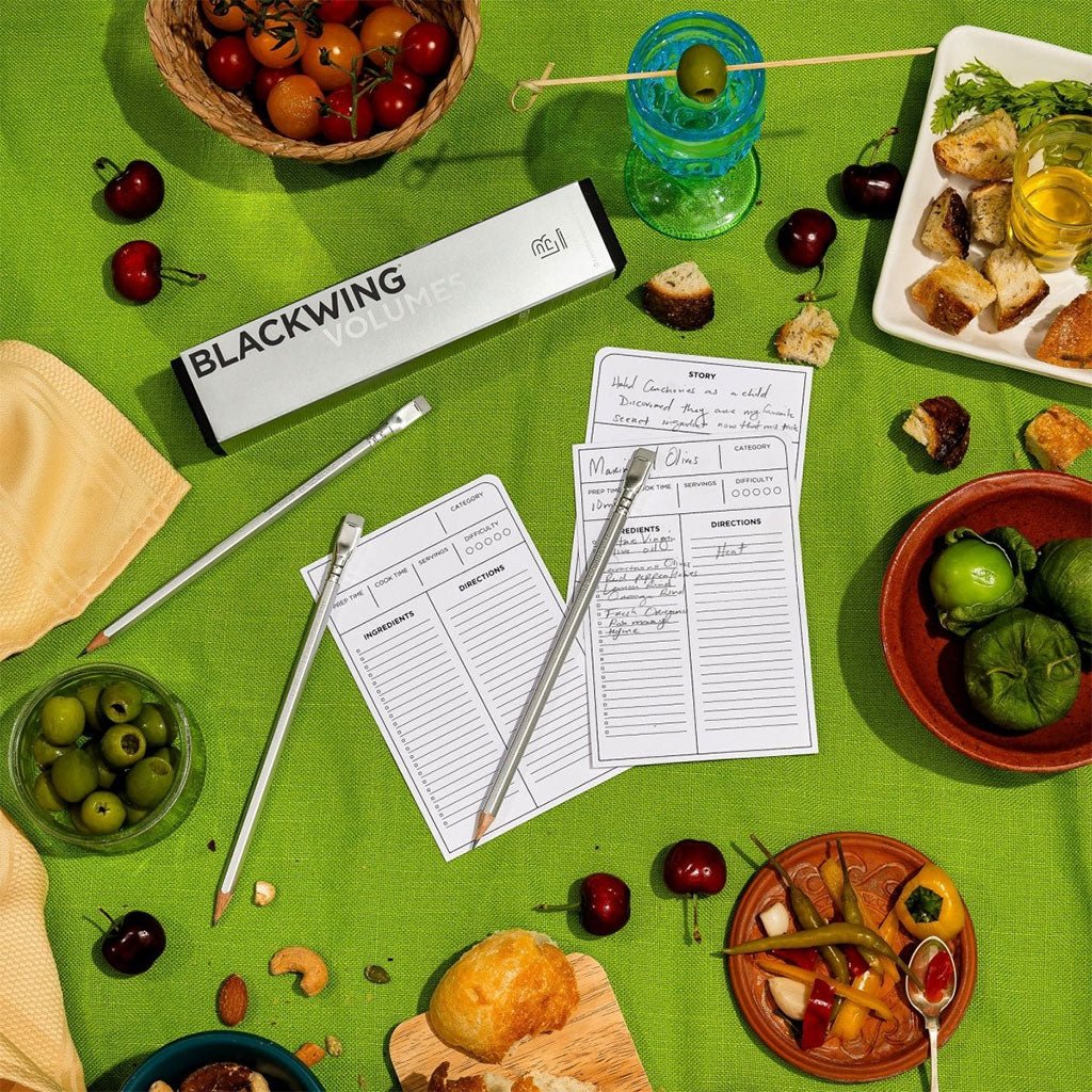 A box of Blackwing Volumes 140 pencils and three pencils sit on a surface covered with a green cloth. They are surrounded by plates of food.