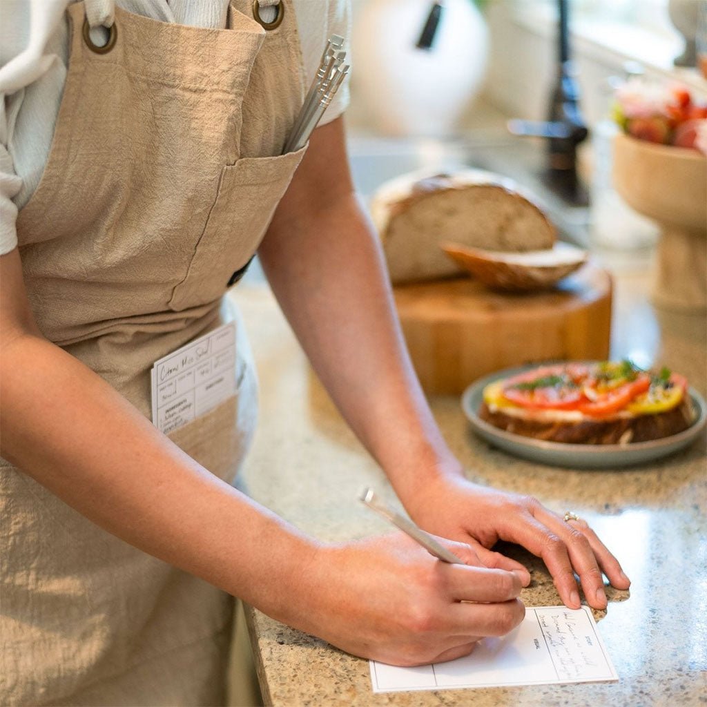 A person writes on a recipe card using a Blackwing Volumes 120 pencil. They are wearing an apron and have three extra pencils tucked in its pocket.