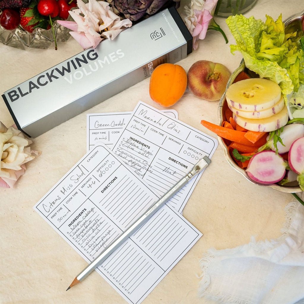 A single Blackwing Volumes 140 pencil sits on a countertop surrounded by recipe cards and cut vegetables and fruit.