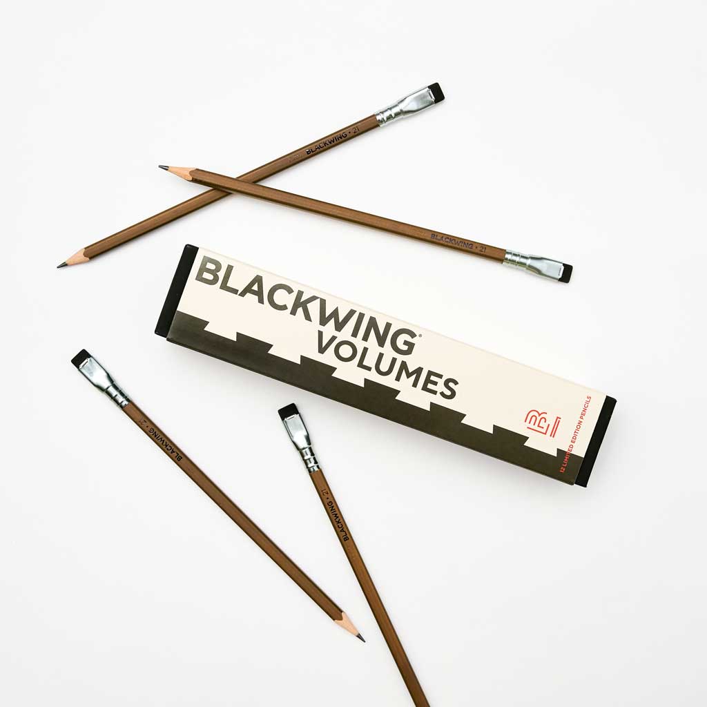 A box of Blackwing Volumes 21 pencils on a white background. Four pencils lay outside the box. They are dark brown with silver ferrules and black erasers.