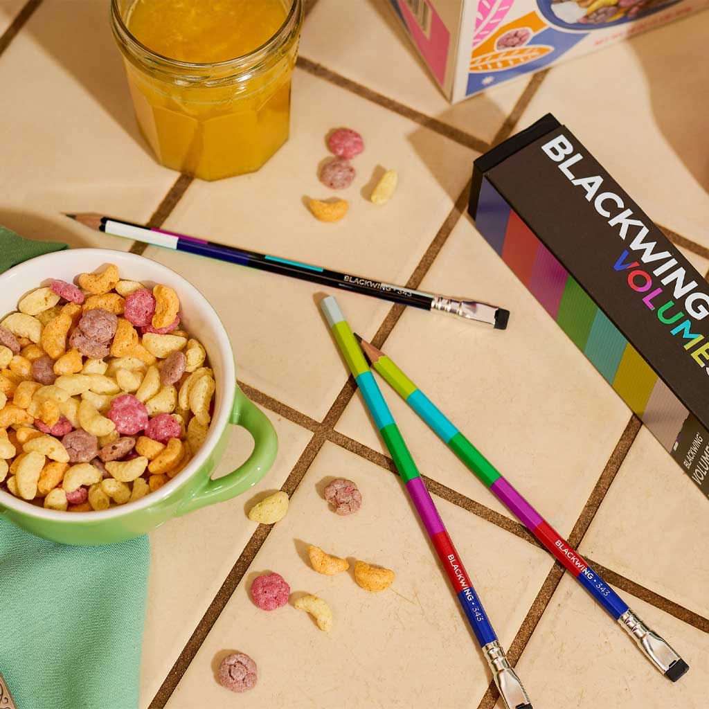 Colorful Blackwing pencils on a tiled surface with a bowl of cereal and a jar of orange juice.