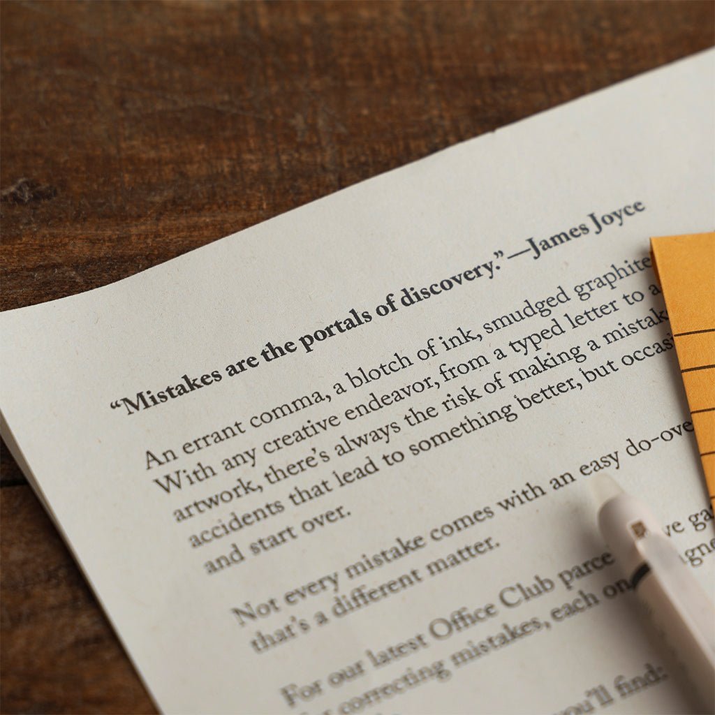 Printed Office Club insert featuring a James Joyce quote about mistakes, resting on a dark wooden surface beside a pen.
