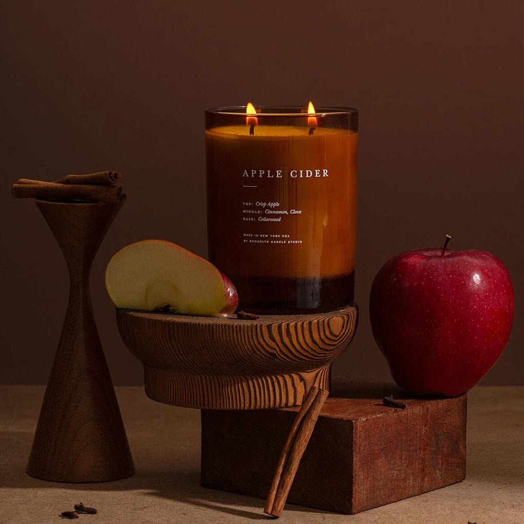 A lit double-wick candle in a clear glass jar labeled 'APPLE CIDER' sits on a wooden stand, styled with a sliced apple, a whole red apple, cinnamon sticks, and dark wood blocks against a warm brown background.