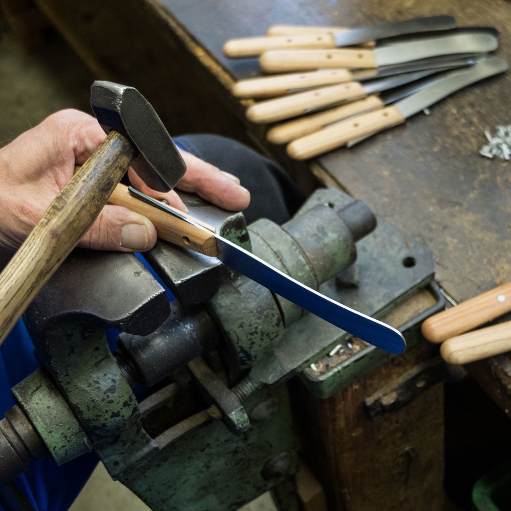 Hand holding a hammer over a blade being inserted into a wooden handle with tools on a workbench
