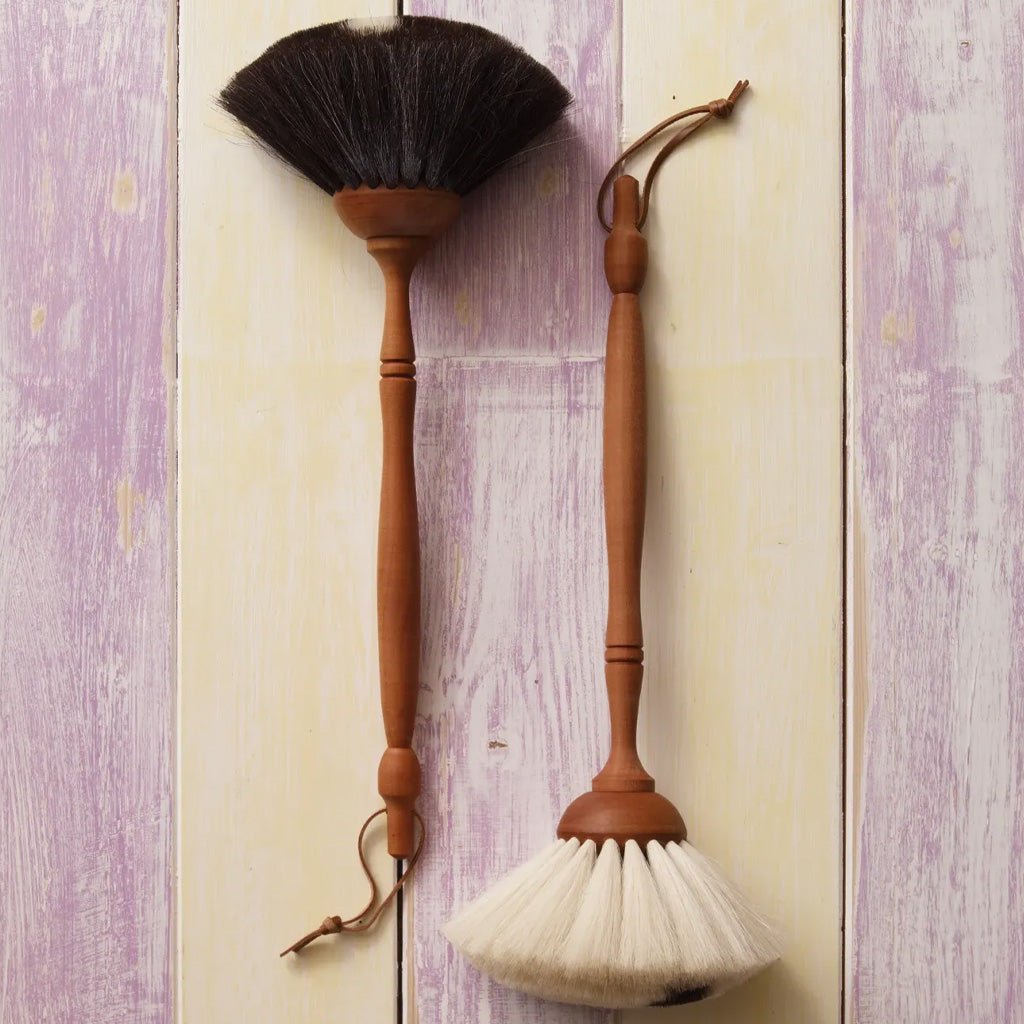 Two wooden dust brushes with long handles hang against a distressed light wood panel background. One brush has dark brown bristles and the other has off-white bristles, both with wood handles and leather ties.