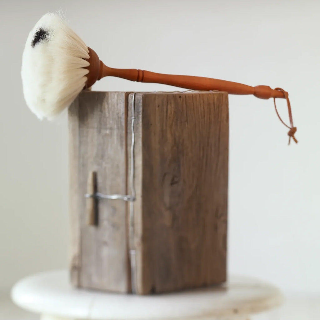 A goat hair dust brush with a light brown wooden handle and white bristles, resting on top of a rustic wooden block.