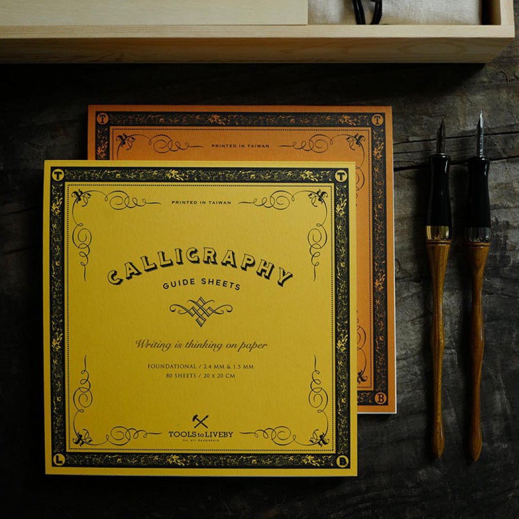 A yellow calligraphy guide sheet with black ornamental border and text is displayed in front of a matching orange book. Two calligraphy brushes with dark handles and gold ferrules lean against a dark textured surface on the right.