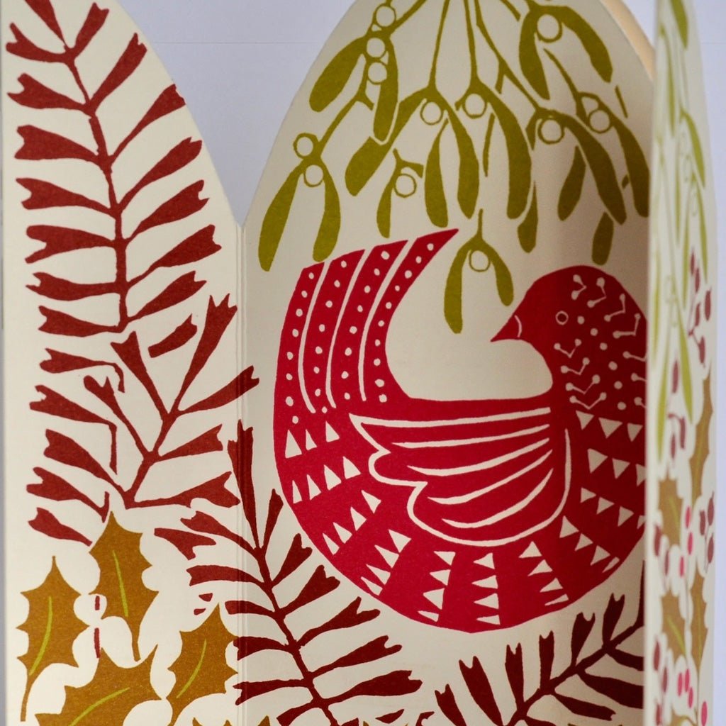 A close-up of unfolded festive paper greeting card featuring a stylized bird in pink and red, surrounded by mistletoe, holly leaves, and berries on a white background.