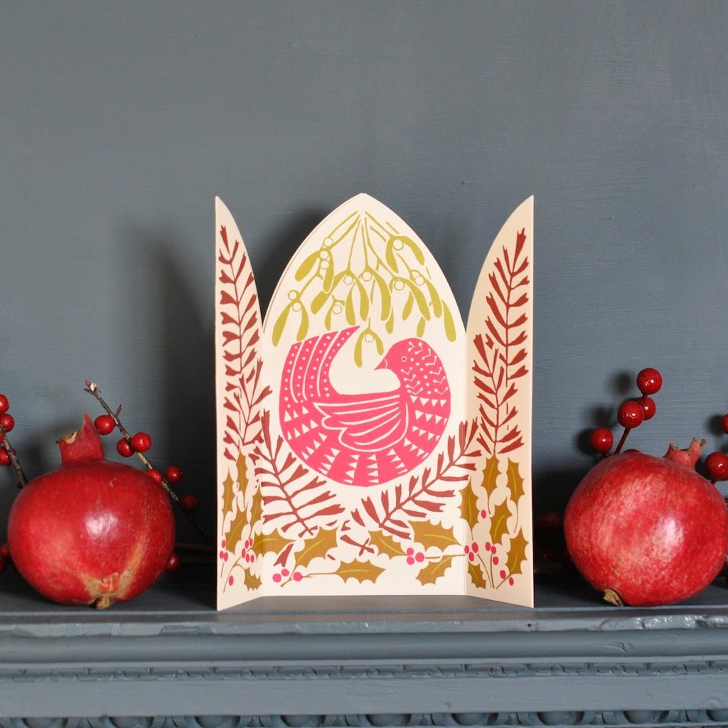A decorative paper card with a Christmas bird and botanical pattern, styled on a gray mantelpiece with red pomegranates and berry sprigs against a dark gray wall.