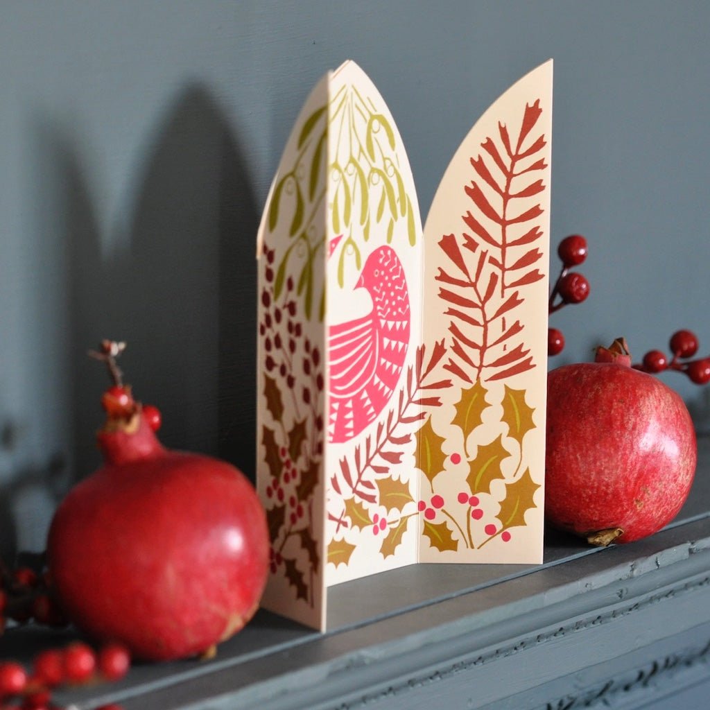 A decorative paper card with a Christmas bird and botanical pattern, styled on a gray mantelpiece with red pomegranates and berry sprigs against a dark gray wall.