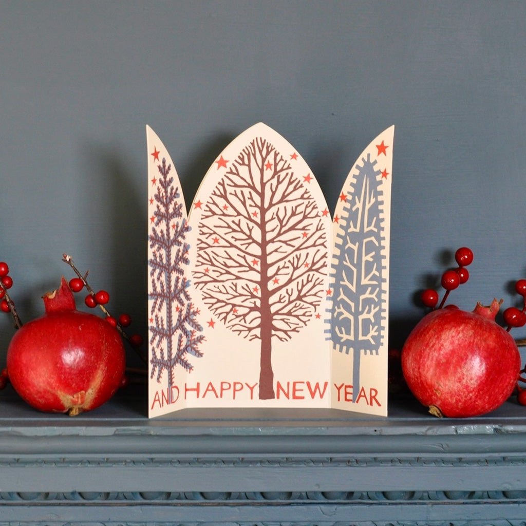 A decorative arched holiday card reading 'AND HAPPY NEW YEAR' with three stylized trees and small red stars, displayed on a gray mantel with pomegranates and red berries against a gray wall.