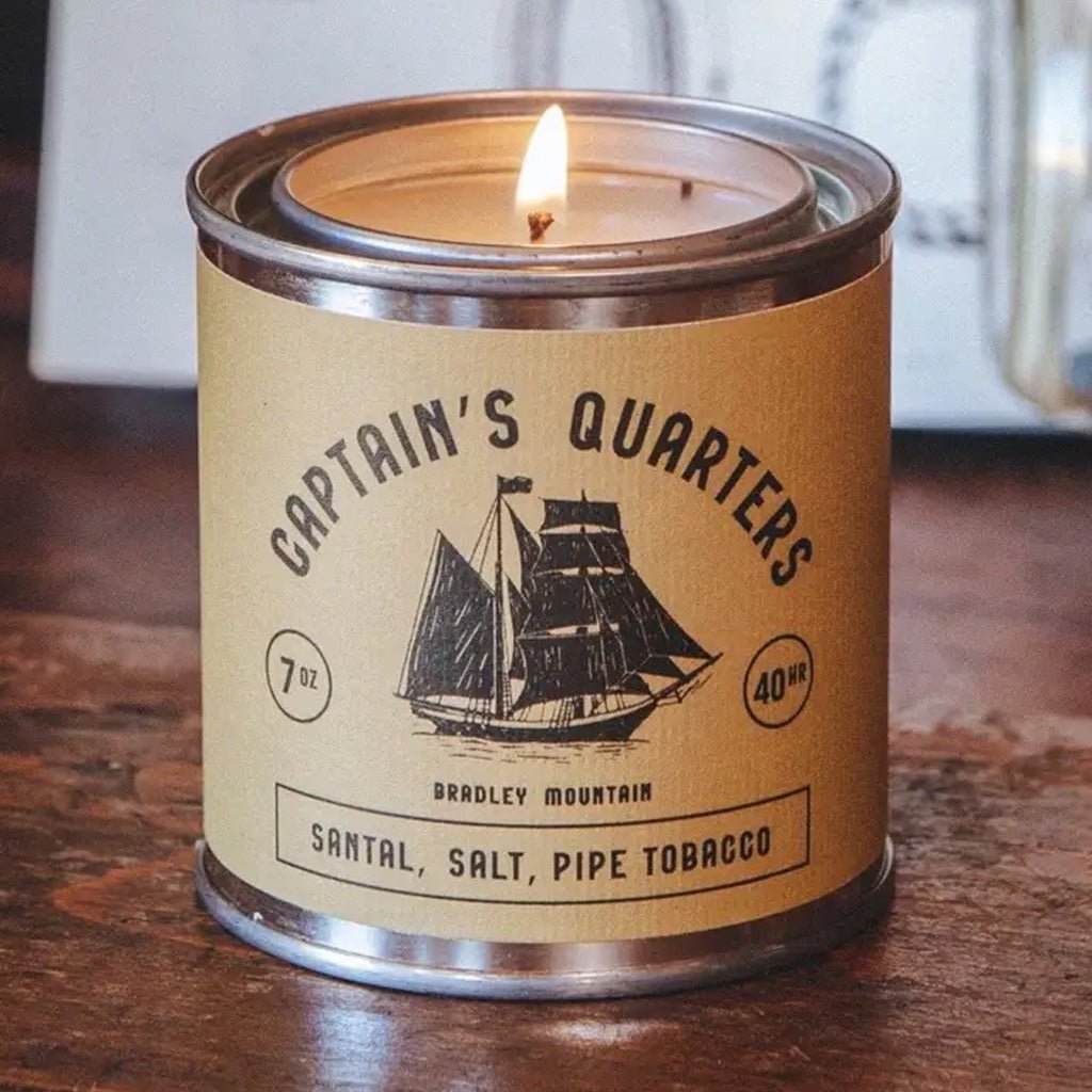 Candle in a tin labeled 'Captain's Quarters' on a wooden surface