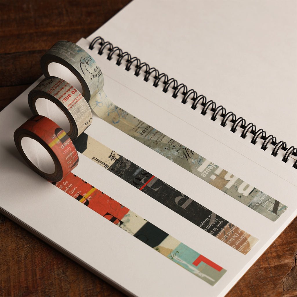 Three rolls of decorative washi tape with vintage-style collage patterns and text, arranged on an open spiral notebook.