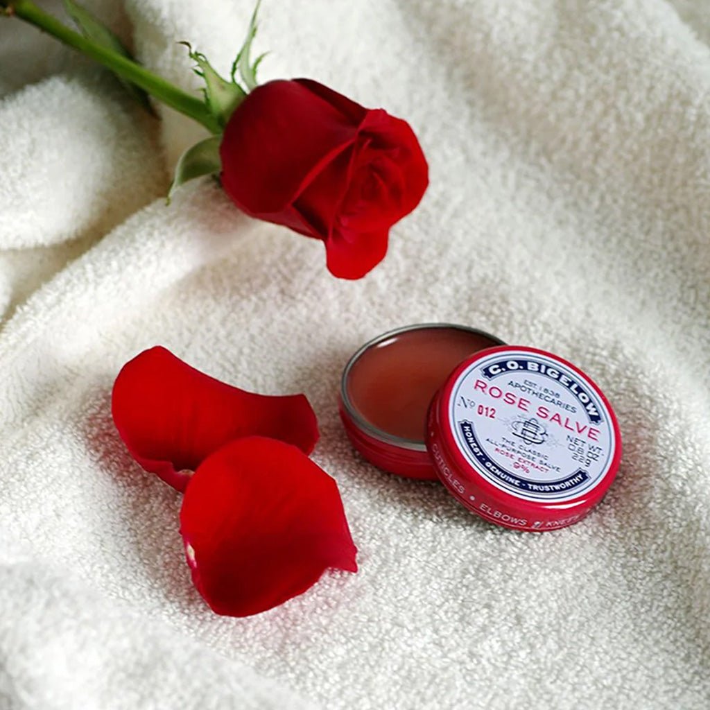 An open tin of rose salve on white towel, next to rose with loose petals.
