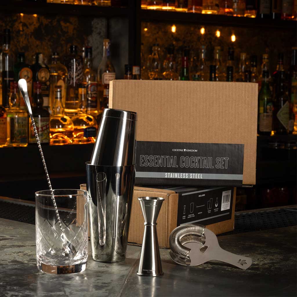 Cocktail shaker, jigger, and strainer on a bar counter with a cocktail box in the background.