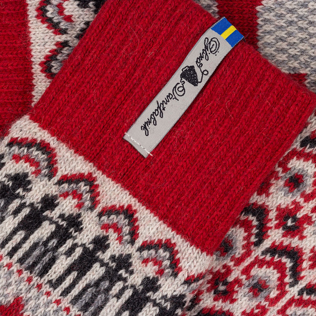 Close-up of a knitted sock cuff in bright red with a white brand label reading 'Öjbro Vantfabrik' and a small blue-yellow accent, over a red, white, and gray patterned knit fabric.