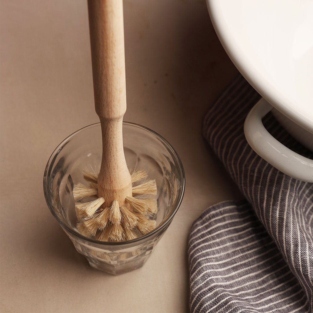 Wooden dish brush in a glass on a beige surface with a striped towel.