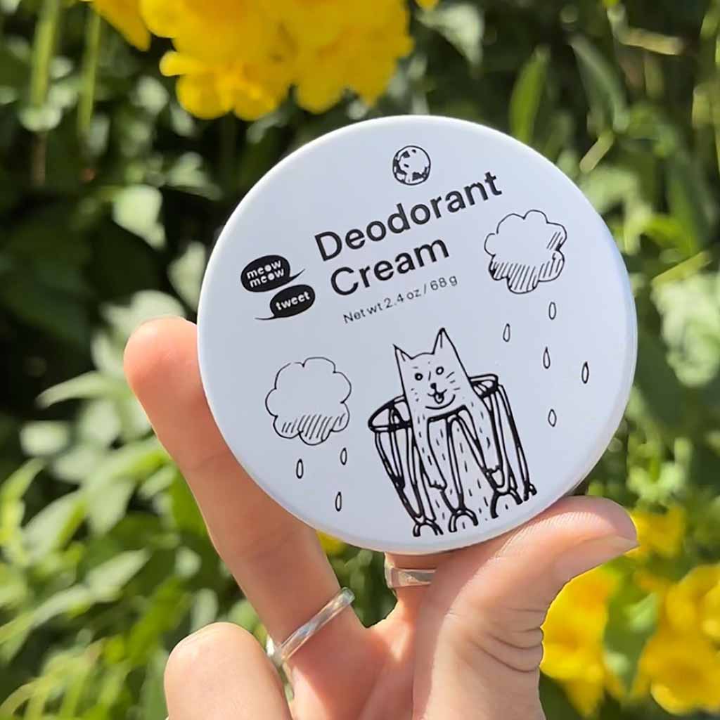 Deodorant cream container with cartoon cat design held by a hand against a natural background with yellow flowers.