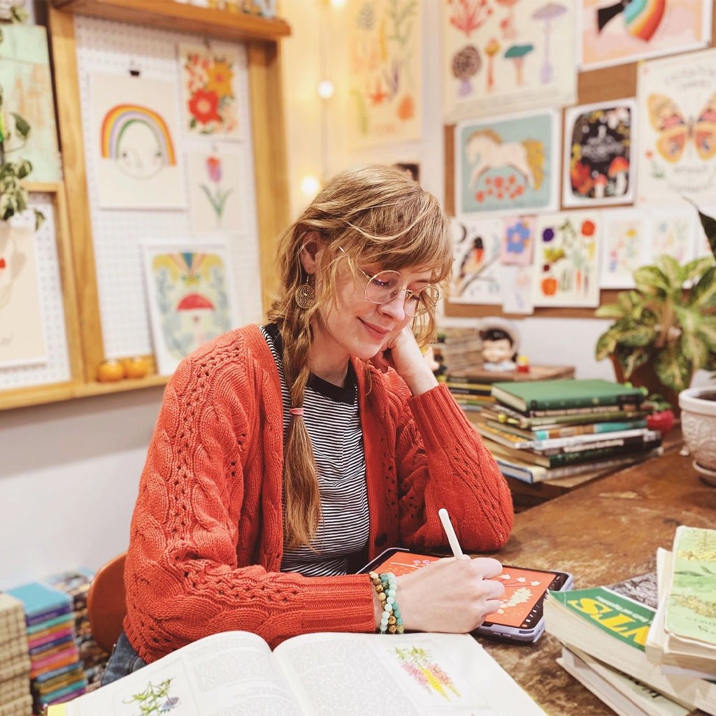 Woman sitting at desk illustrating on a tablet, surrounded by artwork.