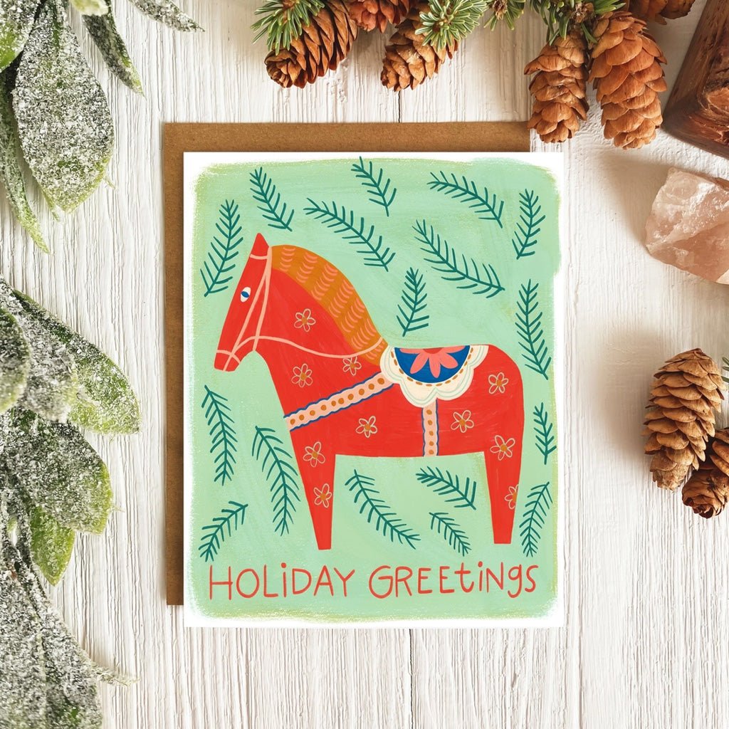 Holiday card with horse on wooden surface surrounded by pinecones.