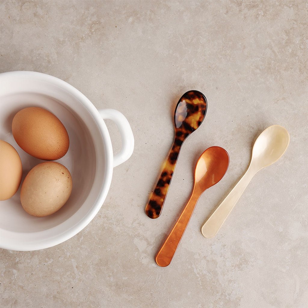 Three spoons with tortoiseshell, cognac brown, and cream finish on a neutral background with eggs in a white bowl.