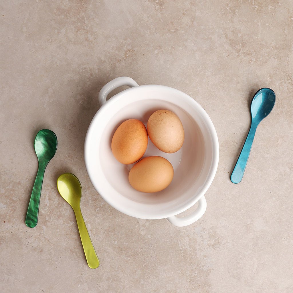 Three eggs in a white bowl with three colorful spoons on a beige surface
