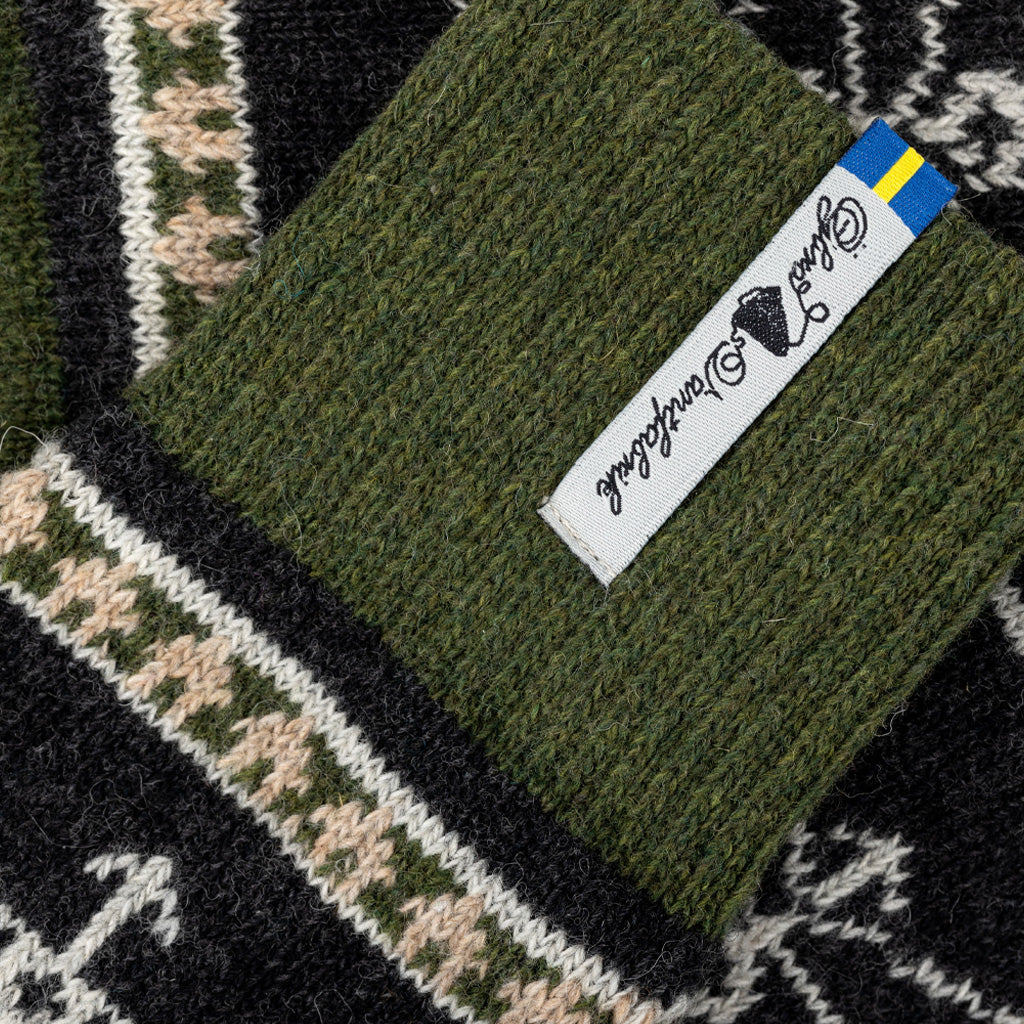 Close-up of a knitted textile with a ribbed olive green cuff featuring a white fabric label that reads 'Ojbro Vantfabrik' and a small blue-yellow flag accent. The cuff is shown over a black, cream, and green patterned knit fabric.