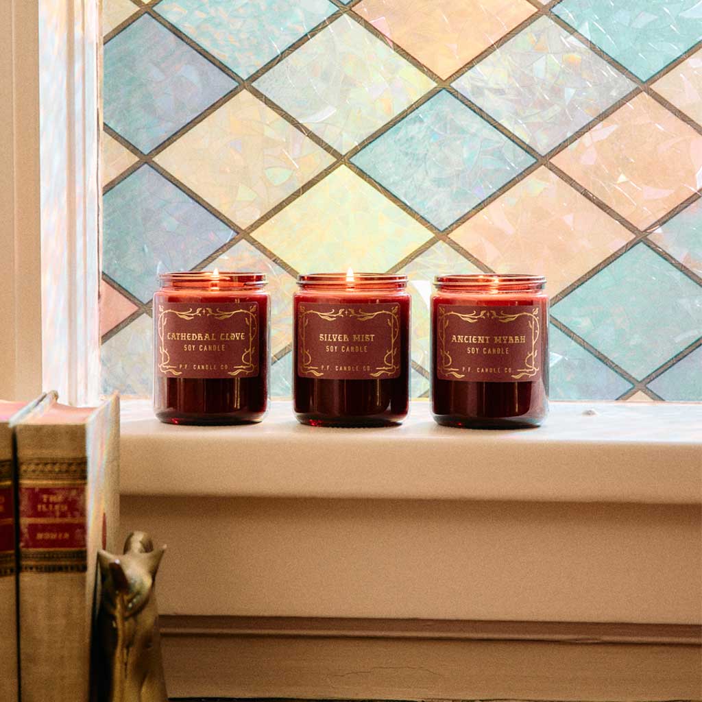 Three candles with labels on a windowsill in front of a colorful tiled window.