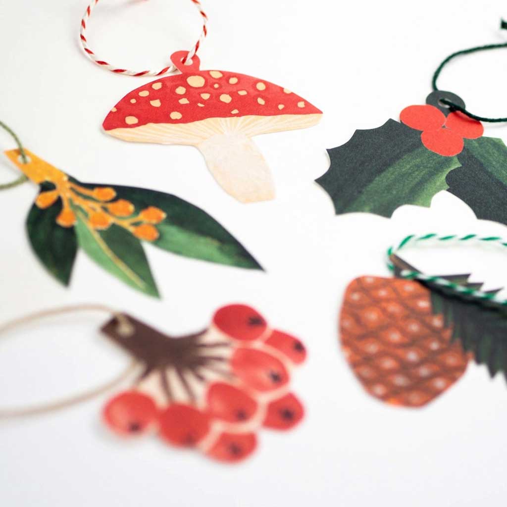 Close up of set of Christmas-themed gift tags including a mushroom, holly, pine cone, and berries on a white background.