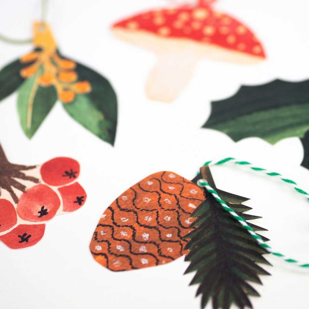 Close up of set of Christmas-themed gift tags including a mushroom, holly, pine cone, and berries on a white background.