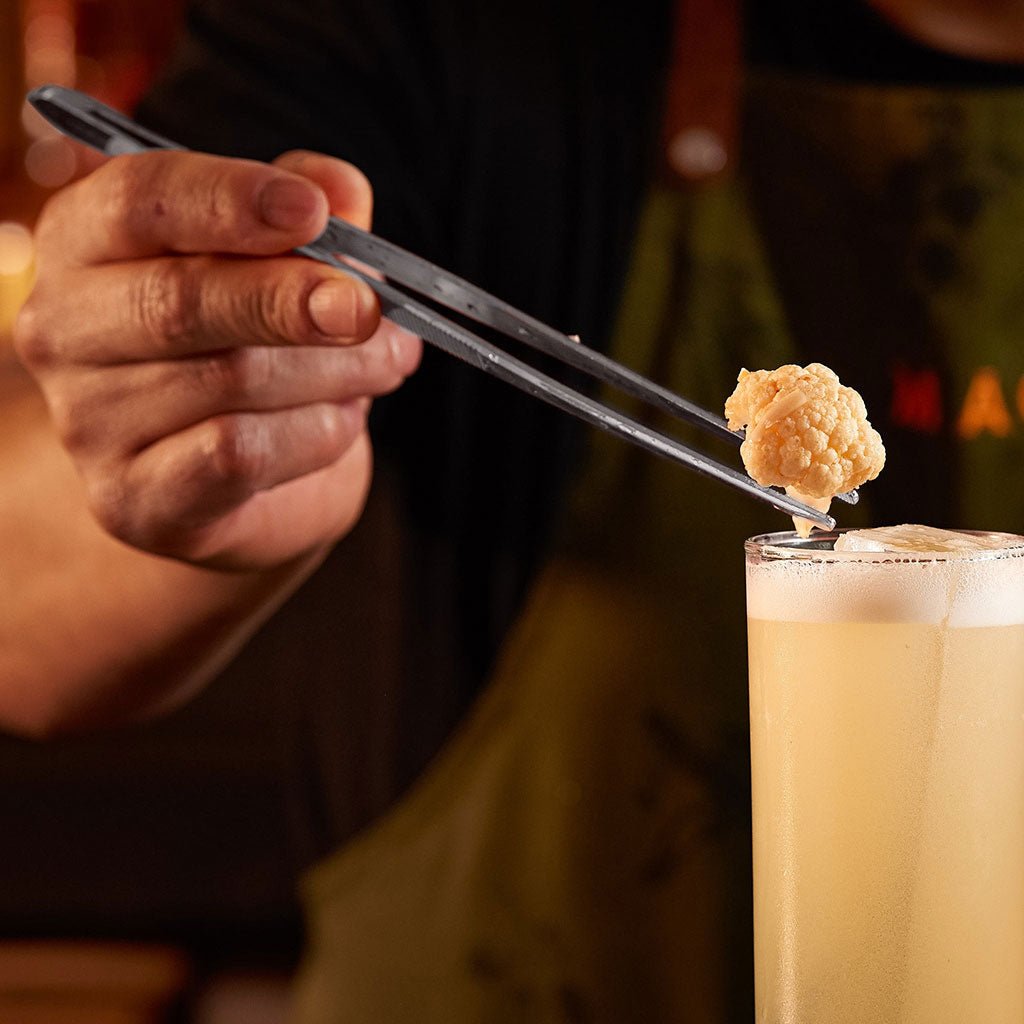 Person using tongs to place a small fried item into a glass of creamy beverage.