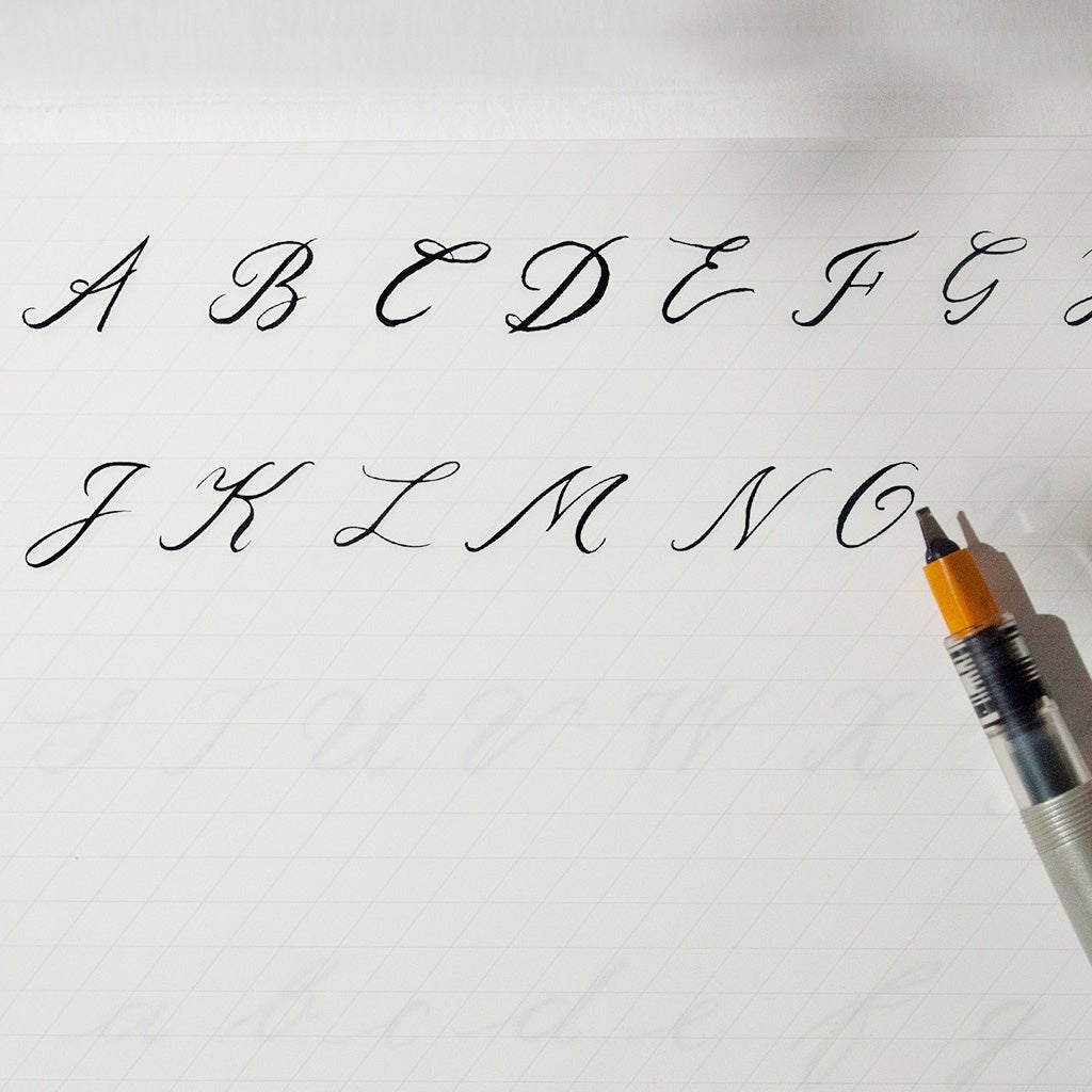 Black calligraphy ink shown with a sample alphabet on white paper, with part of a black and orange calligraphy pen visible in the lower right corner.