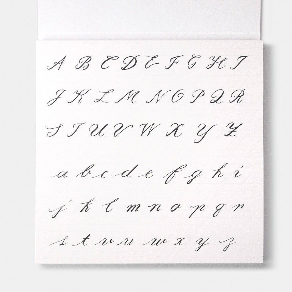 A square white book page showing a cursive alphabet in black ink, with uppercase letters across the top rows and lowercase letters across the bottom rows, photographed against a light neutral background.