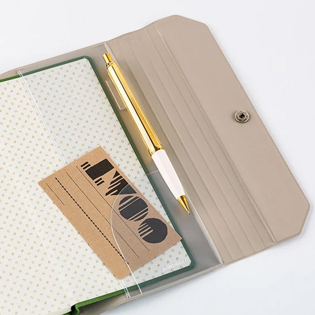 A beige folio-style notebook cover with a snap closure, shown open on a white background. Inside is a white dotted notebook, a gold-and-white pen, and a small brown card with black printed graphics.
