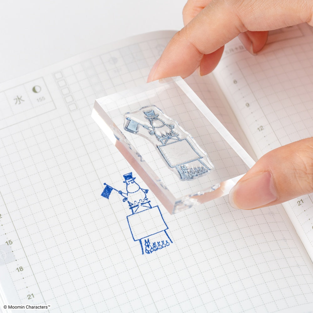 A hand holds a clear acrylic rubber stamp over an open grid notebook, showing a blue inked Moomin character stamp impression below. The stamp itself is transparent with a light blue engraved design.