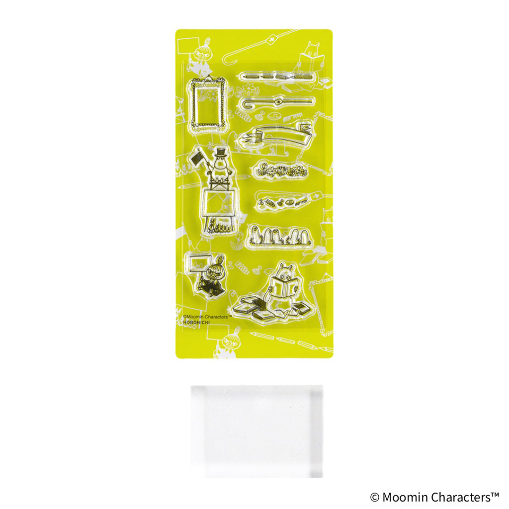 A sheet of clear acrylic stamps featuring various Moomin characters and objects on a bright yellow backing, shown above a clear rectangular stamp block on a white background.