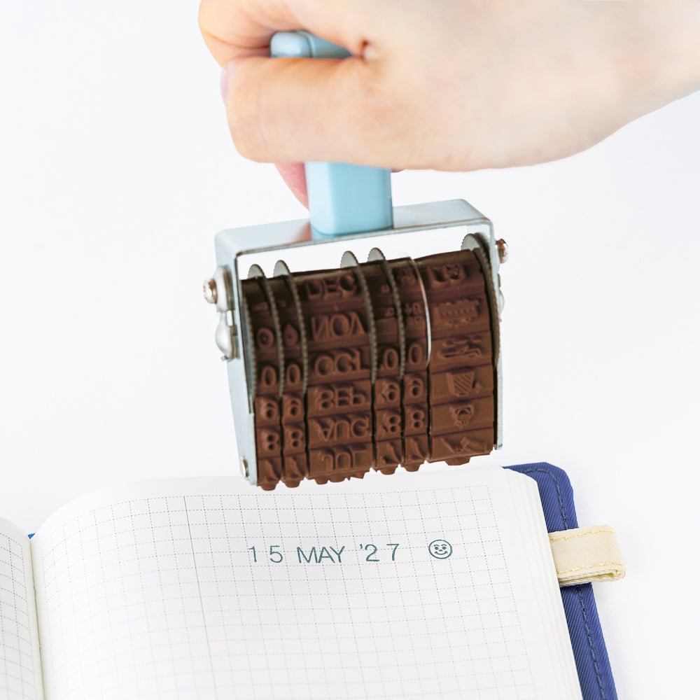 A hand holds a metal rotating stamp over an open grid notebook, showing the engraved date stamping action. The stamp has a light blue handle and a silver metal frame with dark brown engraved letter and number blocks.