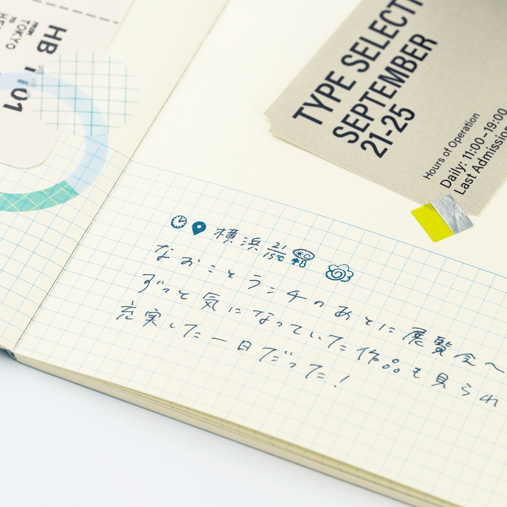 An open cream-colored grid notebook showing handwritten Japanese notes in blue ink, and stamped icons with a beige paper label attached. The notebook is photographed on a clean white background.